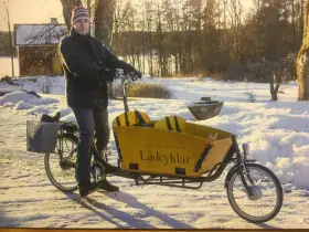 Man med mössa och jacka cyklar på vintern med en gul lastcykel i snöig miljö.